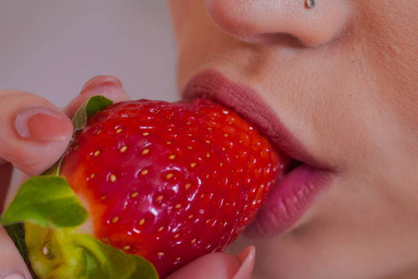 young and beautiful woman eating a very red strawberry while looking mischievously at the camera portrait of seductive woman - Фото, изображение