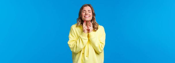 Wishing all well. Hopeful optimistic blond girl in yellow hoodie praying god make wish come true, cross fingers good luck pleading with closed eyes, supplicating, stand blue background. - Foto, afbeelding