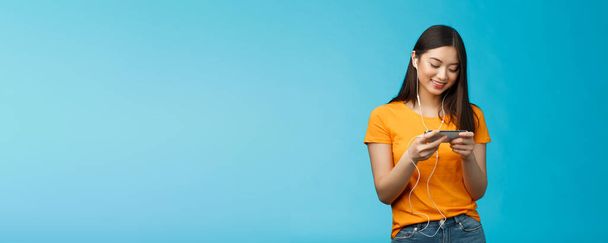 Happy entertained asian cute girl brunette, wear earphones not disturbed people in cafe while playing favorite smartphone game, hold phone horizontal, smiling look enthusiastic cellphone screen. - 写真・画像