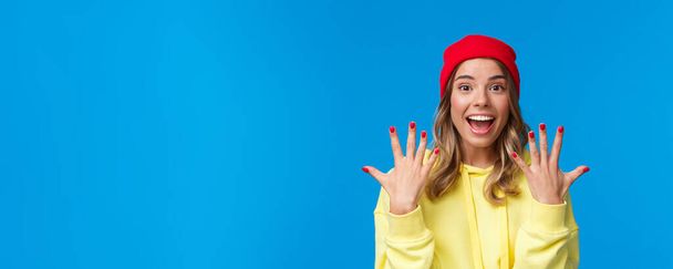 Portrait of amused good-looking blonde hipster girl smiling joyfully, explain something showing number ten, tenth with hands and look camera upbeat, order dozen, stand blue background. - Photo, Image