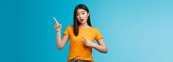 Intrigued curious asian silly tender girl asking question shop assistant opened mouth, questioned interested look, pointing upper right corner puzzled stand blue background. Copy space - 写真・画像