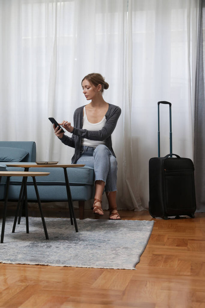 Young woman traveller with smartphone and luggage in hotel room - 写真・画像