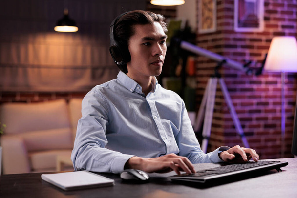 Asian man listening to music on headset and using computer to work remotely, working on business report and research information with headphones and pc. Attending online webinar class. - Φωτογραφία, εικόνα