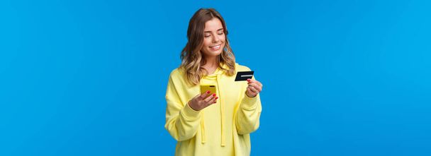 Young caucasian female with blond short hair using mobile phone and insert digit numbers of credit card to pay for her online purchase, shopping in internet store, smiling pleased. - Фото, изображение