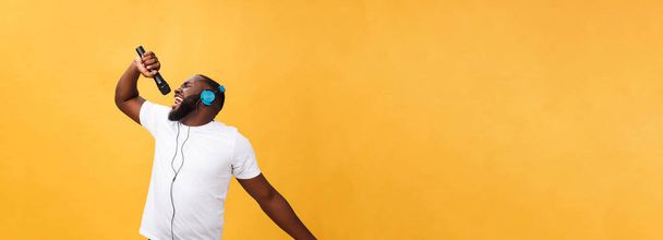 Retrato de alegre chic positivo. hombre africano guapo sosteniendo micrófono y teniendo auriculares en la cabeza escuchando música cantando canción disfrutando de vacaciones de fin de semana aislado sobre fondo amarillo. - Foto, imagen