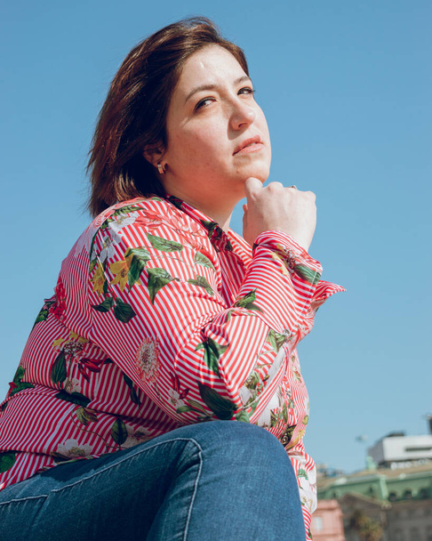 beautiful young Caucasian woman of Argentine ethnicity and plus size, posing crouched with a thoughtful face and a hand on her chin, vertical image for social networks - Photo, Image