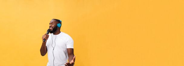 Portret van vrolijke positieve chic. knappe Afrikaanse man met microfoon en koptelefoon op hoofd luisteren muziek zingen lied genieten weekend vakantie geïsoleerd op gele achtergrond. - Foto, afbeelding