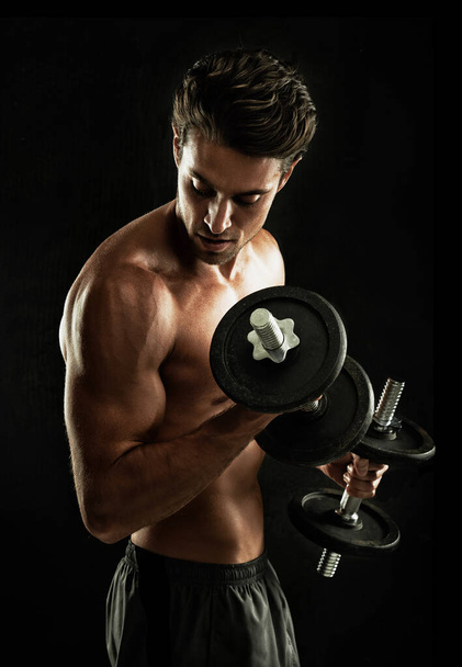 Targeting his biceps. Young man isolated on black performing bicep curls - Foto, afbeelding