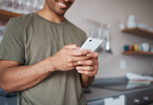 Hands, phone and man texting in a kitchen, relax and smile while reading social media message. Internet, communication and guy planning fun vacation day, streaming and enjoying online app in his home. - Фото, изображение