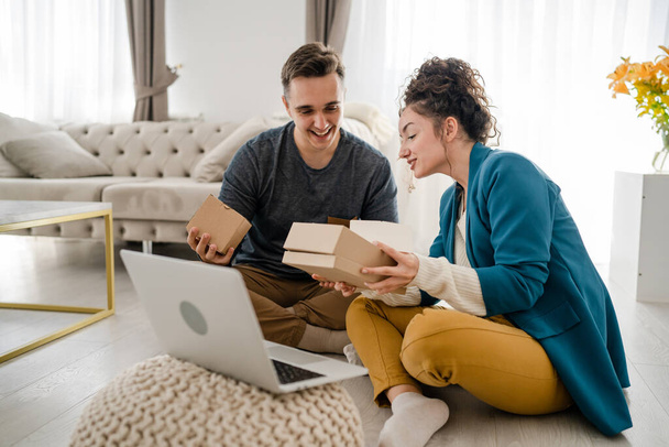 Young caucasian couple woman and man wife and husband or girlfriend and boyfriend opening gifts box presents and read card in front of laptop computer at home having online video call copy space - Photo, Image