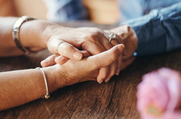 Μακρο, ζευγάρι και ηλικιωμένοι που κρατιούνται χέρι-χέρι στο τραπέζι για υποστήριξη, αγάπη και φροντίδα στο σπίτι. Zoom, ανώτερος και χέρι μαζί για δέσιμο, επέτειο και ειδύλλιο στο γάμο, τη ζωή και τη συνταξιοδότηση. - Φωτογραφία, εικόνα