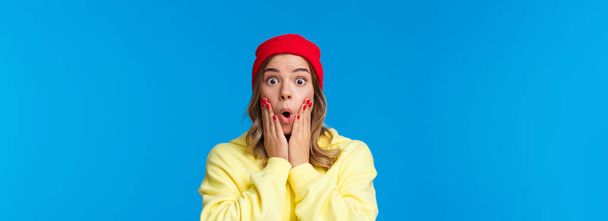 Wow incredible. Impressed and curious good-looking young girl reacting shocked and astonished to interesting awesome news, touch cheeks folding lips stunned, blue background. Copy space - Фото, изображение