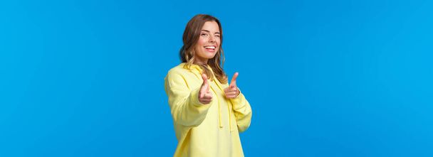 Hey you. Cheeky and carefree cute modern european girl in yellow hoodie, wink and make finger pistols gesture at camera to say hi, informal greeting concept. blue background. - Photo, Image