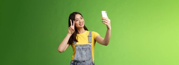 公開かわいいアジアのブルネットの女性は、スマートフォンを保持し、自撮りポーズをとり、電話画面を見て、撮影し、勝利の平和サインを示し、喜んで海外の友人とチャットし、緑の背景に立っている。スペースのコピー - 写真・画像