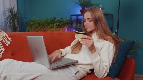 Portrait of caucasian girl using credit bank card and laptop computer while transferring money, purchases online shopping, order food delivery at home. Young woman sitting on couch at home living room - Footage, Video