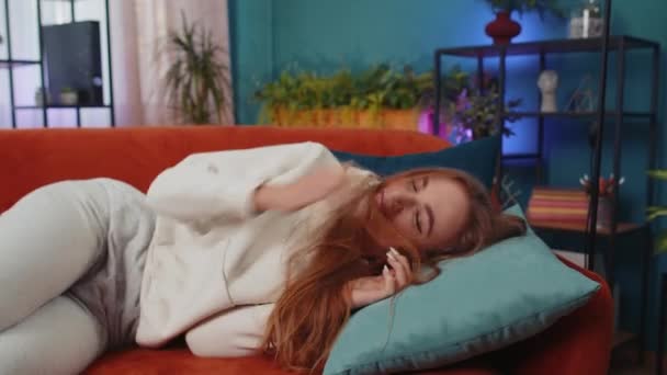 Tired caucasian adult girl lying down in bed taking a rest at home. Carefree young woman napping, falling asleep on comfortable orange sofa with pillows. Closed her eyes enjoy daytime nap alone - Felvétel, videó