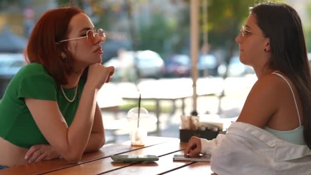 Zwei hübsche Freundinnen unterhalten sich, während sie in einer Bar an der Stadtstraße sitzen - Filmmaterial, Video