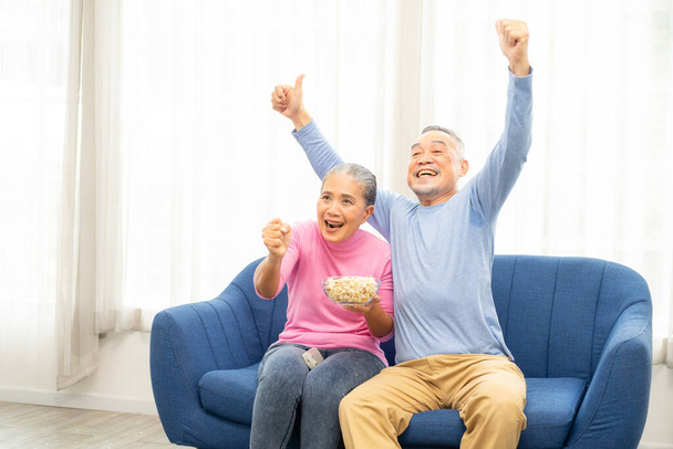 Innoissaan kypsä pari, vanhempi mies ja nainen katsomassa televisiota, vanhempi urheilu fanit juhlivat suosikki joukkue voitto, istuu viihtyisä sohvalla ja syö popcorn välipala kotona, nauttia viikonloppuna. Onnellinen vanhempi. - Valokuva, kuva