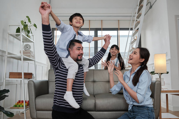 Asian Thai family together, dad plays and teases with children and mum by carrying and holding son on shoulders in home living room, happy leisure times, lovely weekend, wellbeing domestic lifestyle. - Photo, Image