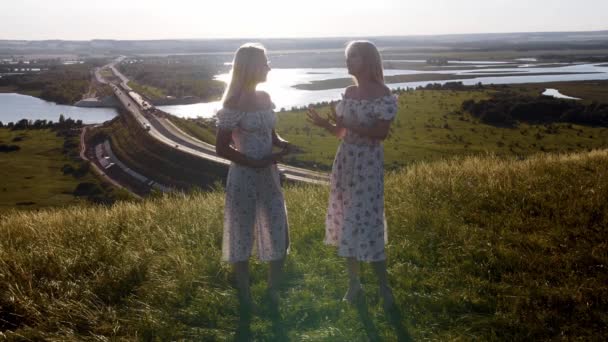Dos jóvenes rubias en vestidos de verano ligeros hablando en una colina. Mid shot - Imágenes, Vídeo