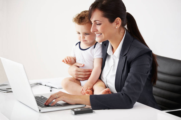 Иногда это возможно иметь все. Улыбающаяся работающая мать, держащая ребенка во время работы над ноутбуком - Фото, изображение