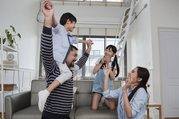 Asian Thai family together, dad plays and teases with children and mum by carrying and holding son on shoulders in home living room, happy leisure times, lovely weekend, wellbeing domestic lifestyle. - Photo, Image