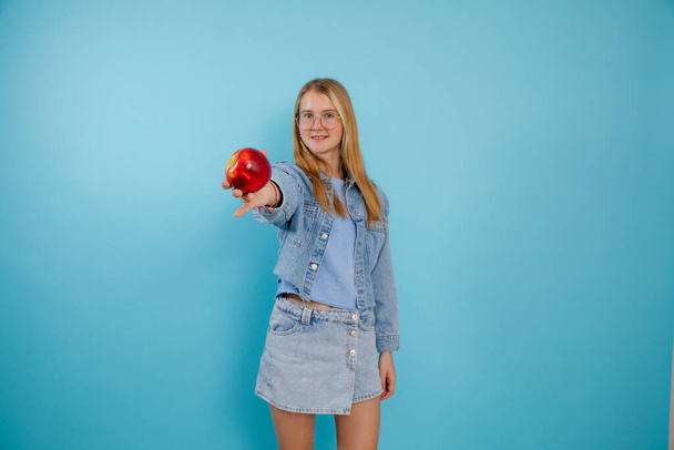Çekici genç kız boş mavi arka planda taze kırmızı elma gösteriyor, bedava fotokopi alanı. Genç bayanın portresi, kollarını uzatmış, olgunlaşmış meyveyi tutuyor. Sağlıklı beslenme, vitaminler. - Fotoğraf, Görsel