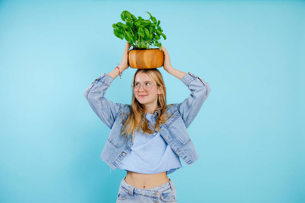 Mooi tienermeisje houden op hoofd groene kamerplant in houten pot op lege blauwe achtergrond, gratis kopieerruimte. Foto van een vrij slanke jonge dame die indoor plant kweekt. Home Bloemkweker, verse planten. - Foto, afbeelding
