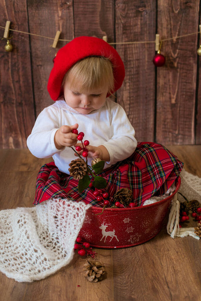 Malá roztomilá dívka v červené kostkované sukni a červený plstěný baret hraje s kužely a vánoční hračky v místnosti zdobené na Vánoce. Vánoce a děti - Fotografie, Obrázek