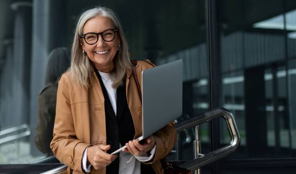 zufriedene ältere Geschäftsfrau mit einem breiten Lächeln und einem Laptop in der Hand vor der Kulisse des Bürozentrums. - Foto, Bild