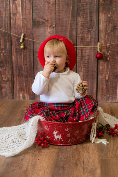 Mała słodka dziewczyna w czerwonej spódnicy w kratę i czerwony beret filcu bawi się szyszki i zabawki świąteczne w pokoju urządzone na Boże Narodzenie. Boże Narodzenie i dzieci - Zdjęcie, obraz