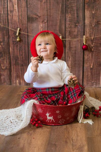 A little cute girl in a red plaid skirt and a red felt beret plays with cones and Christmas toys in a room decorated for Christmas. Christmas and children - Fotografie, Obrázek