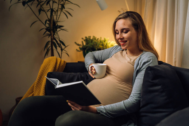 european pregnant woman sit in her living room drinking coffee and reading book - Фото, изображение