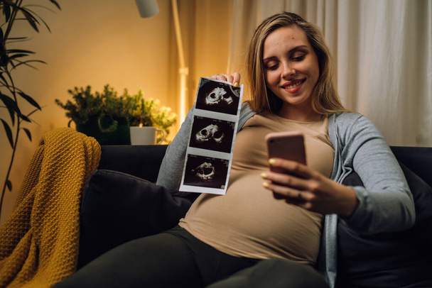 caucasian pregnant woman showing ultrasound photo on video call while sits in her living room - 写真・画像