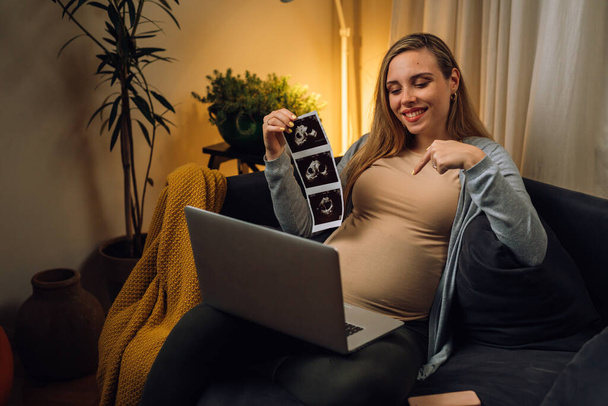 caucasian pregnant woman showing ultrasound photo on video call while sits in her living room - Φωτογραφία, εικόνα