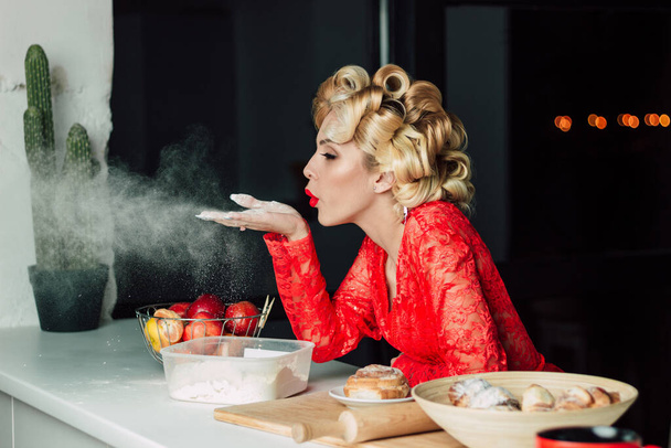 Красивая женщина в красном халате держит муку на руке и дует на нее воздухом. Образ счастливой домохозяйки в красном кружевном халате. Большие кудри на волосах. Правильное питание, диета, приготовление пищи на кухне - Фото, изображение
