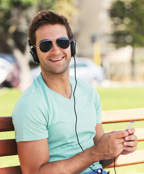Beats on a bench - inside city living. Usmívající se mladý muž se slunečními brýlemi sedící na lavičce v parku a poslouchající hudbu přes sluchátka - Fotografie, Obrázek