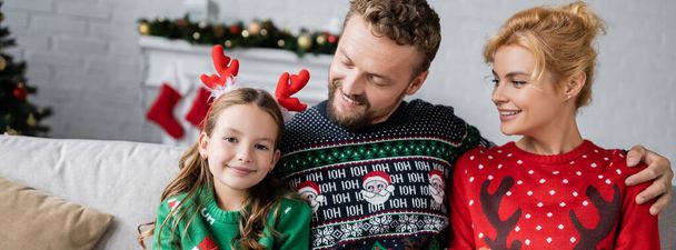 Hymyilevät vanhemmat villapaidoissa katsomassa tytärtä joulun otsanauhalla, bannerilla  - Valokuva, kuva
