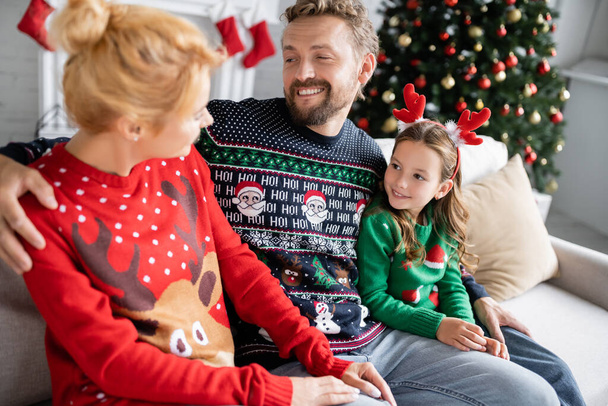 Улыбающийся мужчина обнимает жену и дочь в рождественских свитерах дома  - Фото, изображение