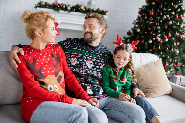 Iloinen mies halaa tytärtä ja vaimoa joulupukissa sohvalla kotona  - Valokuva, kuva