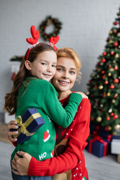 Smiling child hugging mother in sweater holding christmas present at home  - Fotografie, Obrázek