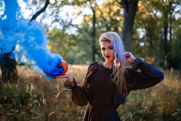 Portrait of beautiful young woman in brown dress holding carved pumpkin with blue smoke coming out of it, in the forest. Halloween party, beautiful woman - Foto, Bild
