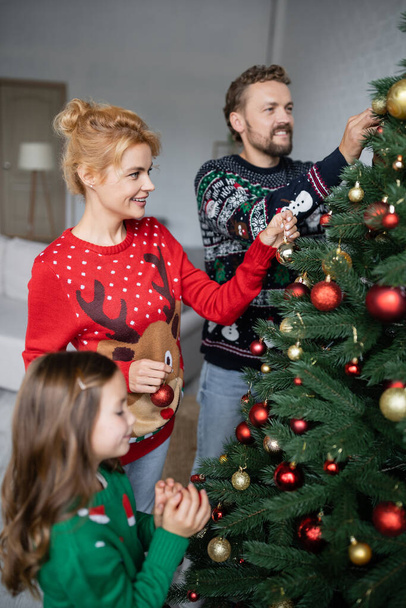 Θετική γυναίκα σε πουλόβερ διακόσμηση χριστουγεννιάτικο δέντρο με την οικογένεια στο σπίτι  - Φωτογραφία, εικόνα