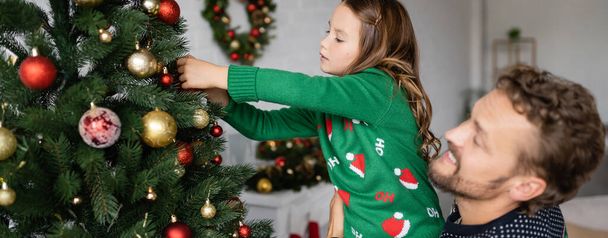 Father holding daughter in sweater decorating christmas tree at home, banner  - Fotografie, Obrázek