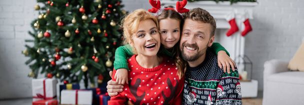 Alegre chica abrazando padre y mamá en suéteres calientes cerca borrosa árbol de Navidad en casa, pancarta  - Foto, imagen