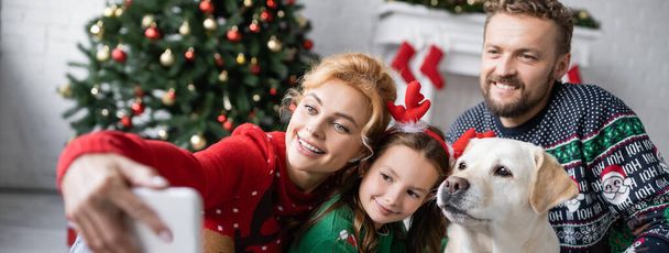Cheerful family with labrador taking selfie on smartphone during new year at home, banner  - Photo, Image