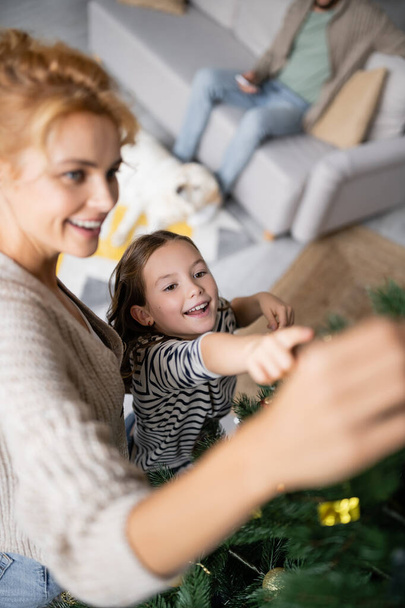 High angle view of girl pointing at christmas tree near blurred mom at home  - Photo, Image