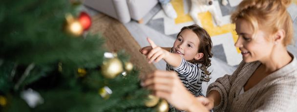 Vue grand angle de fille souriante pointant vers l'arbre de Noël près de maman à la maison, bannière  - Photo, image