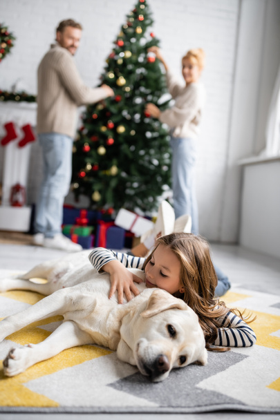 Chica sonriente abrazando a labrador cerca de padres arrugados y árbol de navidad en casa  - Foto, imagen