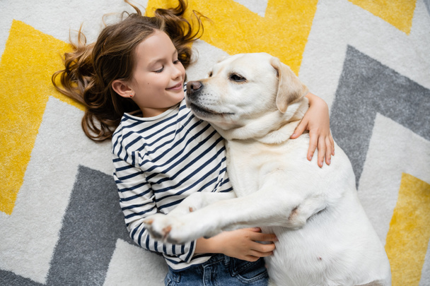 Top view of cheerful kid hugging labrador on floor at home  - Photo, Image